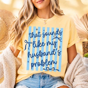 May include: Yellow t-shirt with the text "That sounds like my husband's problem" in blue. The text is within a blue striped rectangle with bow accents. The shirt is a casual, comfortable style.