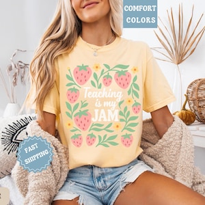 May include: Yellow t-shirt with a strawberry and floral design. The text "Teaching is my JAM" is centered on the front. The shirt is a comfort color and is paired with denim shorts.