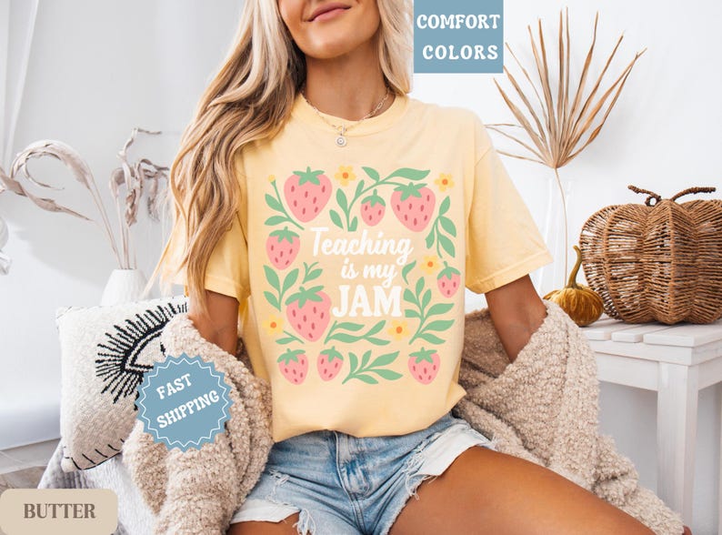 May include: Yellow t-shirt with a strawberry and floral design. The text "Teaching is my JAM" is centered on the front. The shirt is a comfort color and is paired with denim shorts.