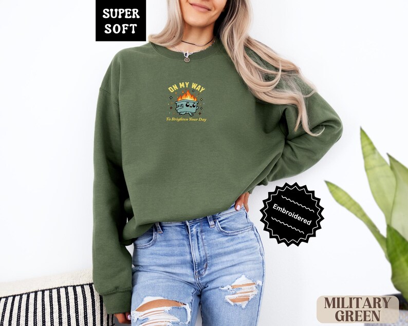 Pu&ograve; includere: Felpa verde militare con grafica ricamata. Il design presenta un'immagine a cartoni animati con il testo "ON MY WAY To Brighten Your Day". La felpa &egrave; etichettata "SUPER SOFT" e "Embroidered".