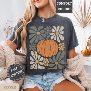 May include: Dark grey t-shirt with a retro design of an orange pumpkin and green and cream daisies. The words "COMFORT COLORS" are printed at the top. The shirt is part of the "PEPPER" collection.