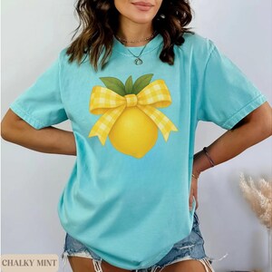 Puede incluir: Una camiseta azul claro con un dise&ntilde;o de lim&oacute;n amarillo y un lazo de cuadros amarillos y blancos. El texto "Chalky Mint" es visible en la esquina inferior izquierda de la imagen.