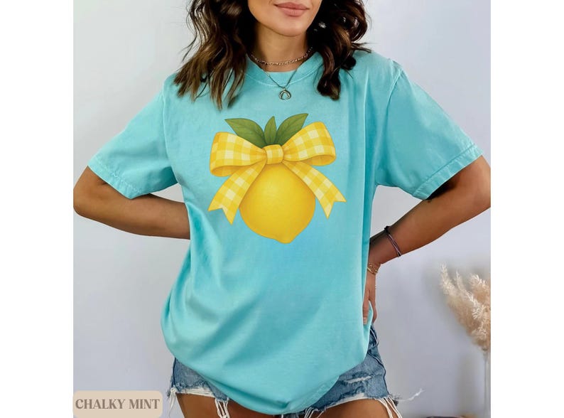 Puede incluir: Una camiseta azul claro con un dise&ntilde;o de lim&oacute;n amarillo y un lazo de cuadros amarillos y blancos. El texto "Chalky Mint" es visible en la esquina inferior izquierda de la imagen.