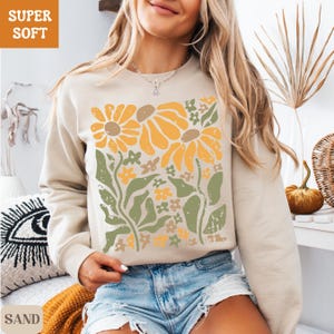 May include: Sand-colored sweatshirt with a floral design in yellow, orange, and green. The graphic features large sunflowers and smaller flowers with leaves. The text "SUPER SOFT" is in the upper left corner, and "SAND" is in the lower left corner.