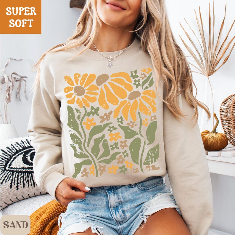 May include: Sand-colored sweatshirt with a floral design in yellow, orange, and green. The graphic features large sunflowers and smaller flowers with leaves. The text "SUPER SOFT" is in the upper left corner, and "SAND" is in the lower left corner.