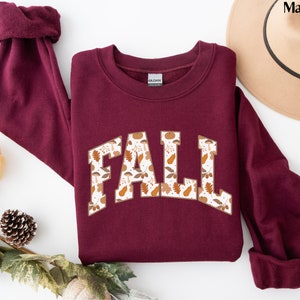 K&ouml;nnte beinhalten: Ein maroonfarbener Pullover mit einem wei&szlig;en "FALL"-Motiv. Das Motiv ist mit einem Muster aus Herbstbl&auml;ttern und K&uuml;rbissen gef&uuml;llt.