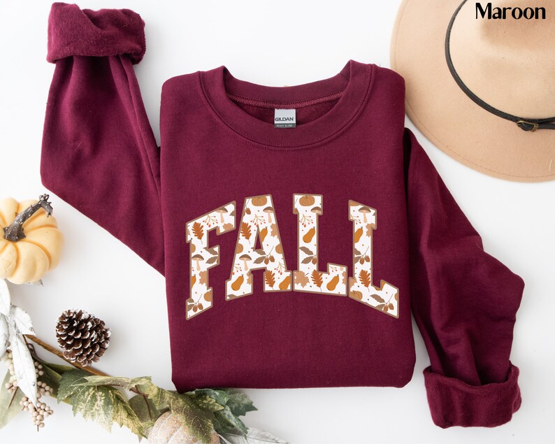 K&ouml;nnte beinhalten: Ein maroonfarbener Pullover mit einem wei&szlig;en "FALL"-Motiv. Das Motiv ist mit einem Muster aus Herbstbl&auml;ttern und K&uuml;rbissen gef&uuml;llt.