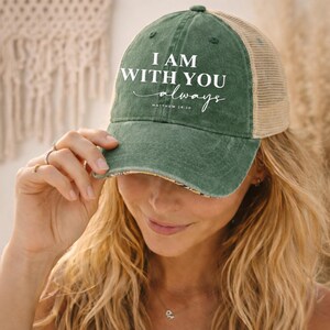May include: Green and tan trucker hat with the text "I AM WITH YOU always" in white. The hat has a distressed look and a curved brim. The text "Matthew 28:20" is also printed on the hat.