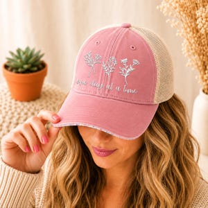 May include: A dusty rose baseball cap with a beige mesh back. The cap features embroidered white floral designs and the phrase "one day at a time" in a script font. The brim has a floral pattern.