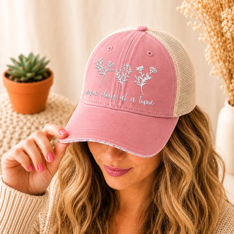 May include: A dusty rose baseball cap with a beige mesh back. The cap features embroidered white floral designs and the phrase "one day at a time" in a script font. The brim has a floral pattern.