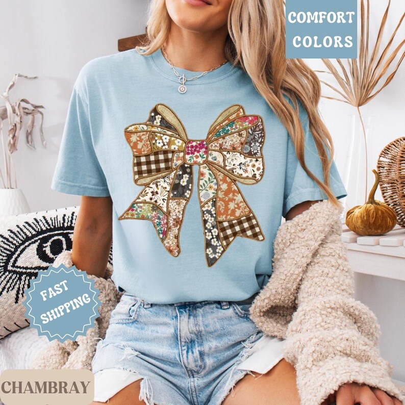 May include: A light blue t-shirt featuring a large, patchwork bow design. The bow incorporates various patterns, including floral, gingham, and solid colors. The words "COMFORT COLORS" are printed in a white box. The shirt is labeled "CHAMBRAY" and "FAST SHIPPING".