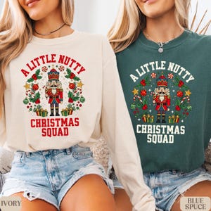 May include: Two long-sleeved shirts, one ivory and one teal, each featuring a festive Christmas design. The design includes a nutcracker, the text "A Little Nutty," and "Christmas Squad" in red. The shirts are paired with denim shorts.