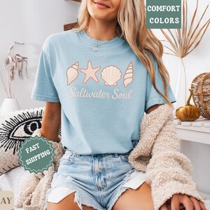 May include: Light blue t-shirt with a seashell design and the words "Saltwater Soul". The shirt is made of chambray fabric and is paired with denim shorts. The image also includes a cream-colored sweater.