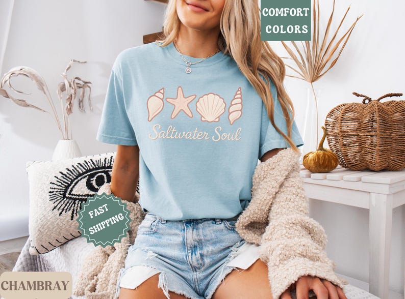 May include: Light blue t-shirt with a seashell design and the words "Saltwater Soul". The shirt is made of chambray fabric and is paired with denim shorts. The image also includes a cream-colored sweater.