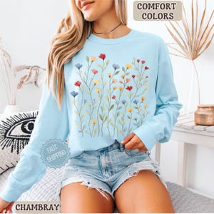 May include: A light blue long-sleeve sweatshirt with a floral design. The design features colorful flowers in red, yellow, and blue. The words "COMFORT COLORS" are in a tan box in the upper right corner. The word "CHAMBRAY" is at the bottom.
