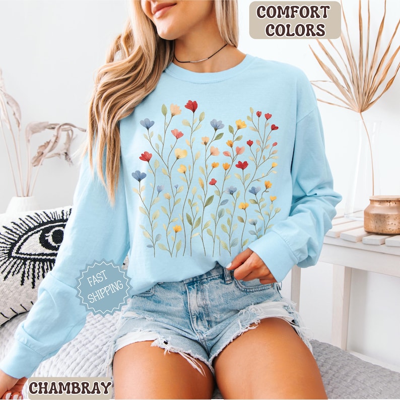 May include: A light blue long-sleeve sweatshirt with a floral design. The design features colorful flowers in red, yellow, and blue. The words "COMFORT COLORS" are in a tan box in the upper right corner. The word "CHAMBRAY" is at the bottom.