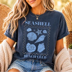May include: Navy blue t-shirt with a light blue seashell graphic and the words "Seashell Beach Club". The graphic features various seashell illustrations within a rectangular frame. The shirt has rolled-up sleeves.