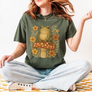 May include: A green frog perched on a mushroom with red and white spots. The mushroom is surrounded by yellow flowers and green leaves. The frog is wearing a green shirt with a floral pattern.