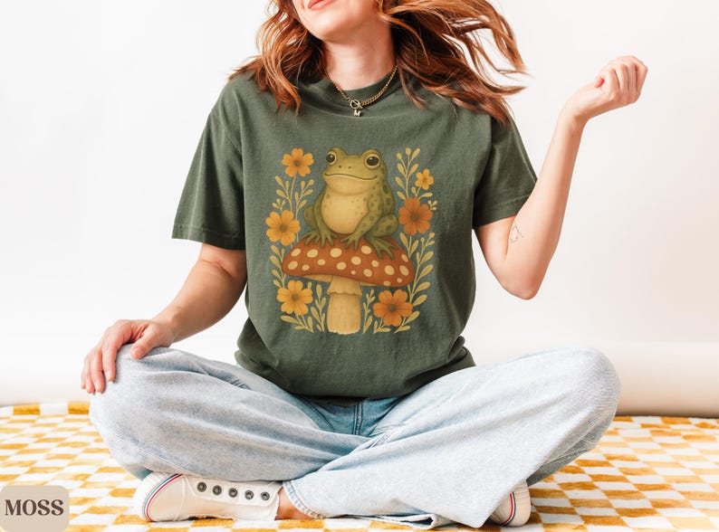 May include: A green frog perched on a mushroom with red and white spots. The mushroom is surrounded by yellow flowers and green leaves. The frog is wearing a green shirt with a floral pattern.