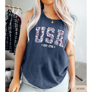 K&ouml;nnte beinhalten: Ein marineblaues Tanktop mit dem Wort "USA" in einem floralen Muster. Unter dem Wort "USA" steht der Text "EST. 1776" mit Sternsymbolen. Das Tanktop ist aus Denim-Material.