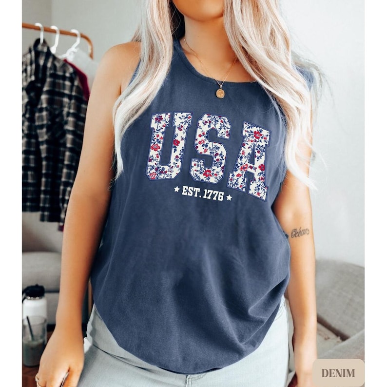 K&ouml;nnte beinhalten: Ein marineblaues Tanktop mit dem Wort "USA" in einem floralen Muster. Unter dem Wort "USA" steht der Text "EST. 1776" mit Sternsymbolen. Das Tanktop ist aus Denim-Material.