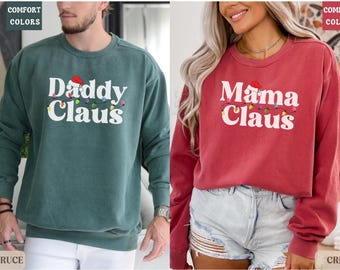 May include: Two Christmas themed sweatshirts, one green with "Daddy Claus" and the other red with "Mama Claus" printed on them. Both sweatshirts feature a Santa hat and string lights.