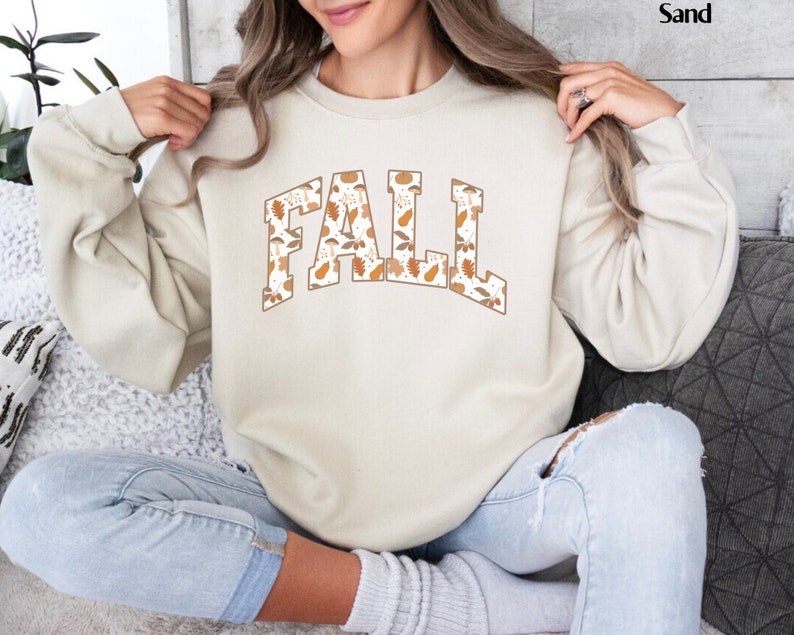 May include: A sand-colored sweatshirt with the word "FALL" in a collegiate-style font. The letters are filled with a pattern of fall leaves and gourds in shades of brown and orange. The sweatshirt has long sleeves and a crew neck.