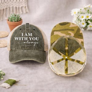 May include: Olive green trucker hat with the text "I AM WITH YOU always" in white script. The hat features a mesh back and a camouflage pattern on the underside of the brim. The text "Matthew 28:20" is below the main text.