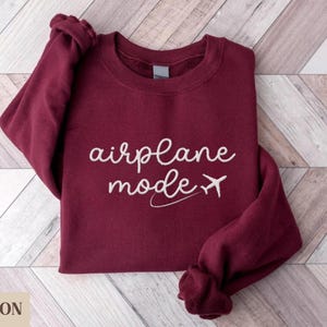 Puede incluir: Una sudadera color burdeos con el texto "airplane mode" y un avi&oacute;n blanco.