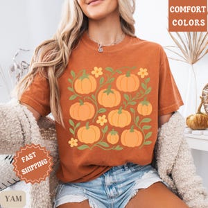 May include: Orange t-shirt featuring a pumpkin design with green vines and yellow flowers. The shirt has the words "COMFORT COLORS" printed on the upper right side. The shirt is a fall-themed design.