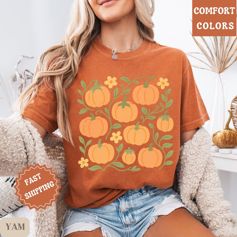 May include: Orange t-shirt featuring a pumpkin design with green vines and yellow flowers. The shirt has the words "COMFORT COLORS" printed on the upper right side. The shirt is a fall-themed design.