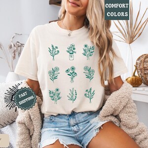 May include: A woman wearing a white t-shirt with a green floral design. The shirt has the text "COMFORT COLORS" on the top right corner. The woman is wearing denim shorts and a white fuzzy jacket. The background is a light brown wall with a wicker basket and a plant.