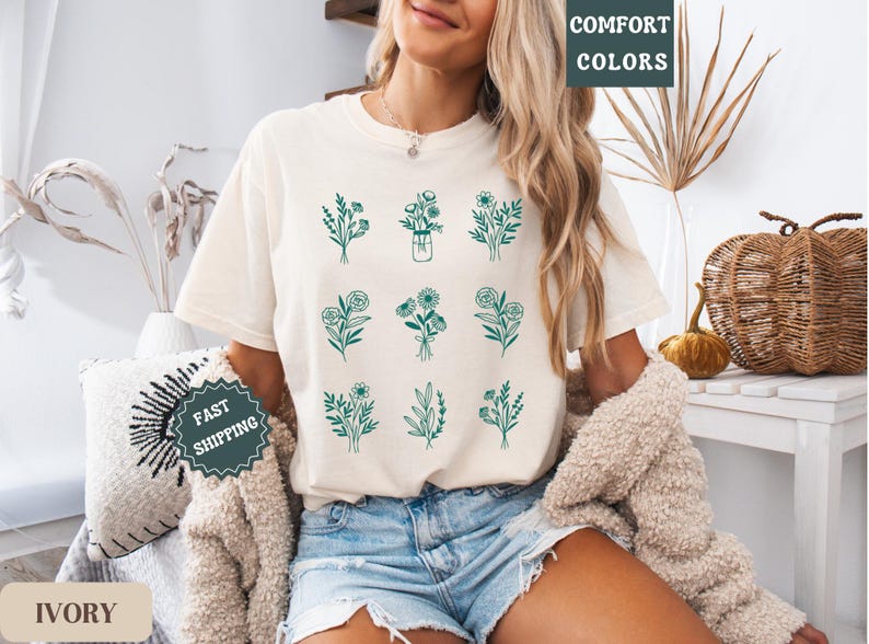 May include: A woman wearing a white t-shirt with a green floral design. The shirt has the text "COMFORT COLORS" on the top right corner. The woman is wearing denim shorts and a white fuzzy jacket. The background is a light brown wall with a wicker basket and a plant.