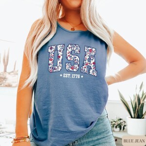 May include: A blue tank top with the word "USA" in a floral design. The text "EST. 1776" is below the word "USA". The tank top is a solid blue color.