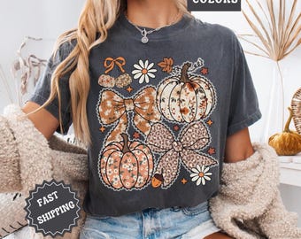 May include: Dark gray t-shirt featuring a fall-themed graphic with pumpkins, bows, and floral accents. The design includes the text "COMFORT COLORS" and "FAST SHIPPING". The shirt is a relaxed fit.