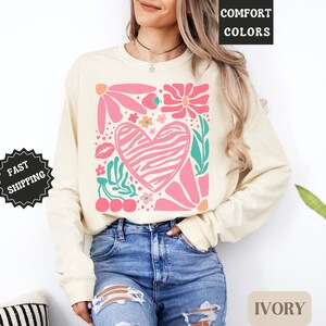 May include: Ivory-colored long-sleeve sweatshirt with a colorful graphic. The graphic features a pink zebra-striped heart, flowers, leaves, cherries, and a strawberry. The words "COMFORT COLORS" are in the upper right corner.