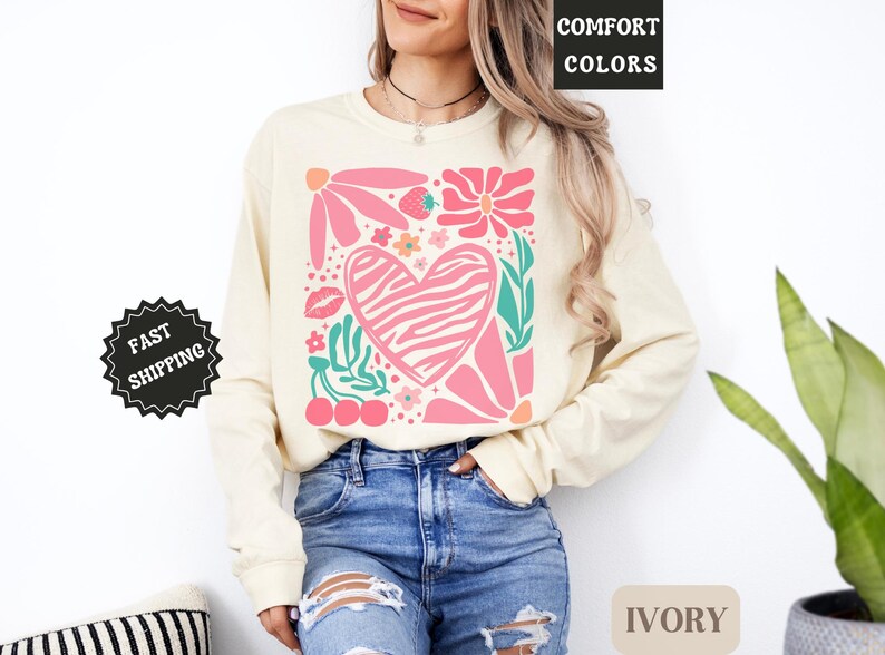May include: Ivory-colored long-sleeve sweatshirt with a colorful graphic. The graphic features a pink zebra-striped heart, flowers, leaves, cherries, and a strawberry. The words "COMFORT COLORS" are in the upper right corner.