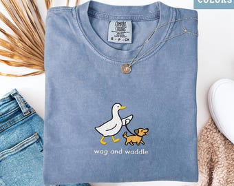 May include: Blue jean colored t-shirt with a graphic of a white duck walking a small brown dog on a leash. The text "wag and waddle" is below the graphic. The shirt has a Comfort Colors tag.