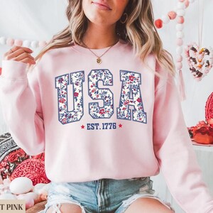 Peut inclure: Sweat-shirt rose clair avec le mot "USA" dans un motif floral. Le texte "EST. 1776" est en dessous du mot "USA". Le sweat-shirt est de couleur rose clair.