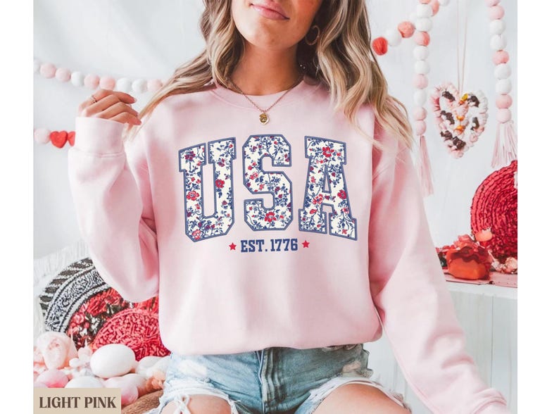 Peut inclure: Sweat-shirt rose clair avec le mot "USA" dans un motif floral. Le texte "EST. 1776" est en dessous du mot "USA". Le sweat-shirt est de couleur rose clair.
