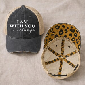 May include: A black and tan trucker hat with the text "I AM WITH YOU always" in white script. The hat has a leopard print lining and a mesh back. The hat is a baseball cap style.