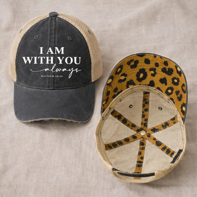 May include: A black and tan trucker hat with the text "I AM WITH YOU always" in white script. The hat has a leopard print lining and a mesh back. The hat is a baseball cap style.