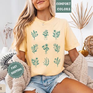 May include: A yellow t-shirt with a floral design. The shirt has a crew neck and short sleeves. The design features nine different floral arrangements in a green color. The text "COMFORT COLORS" is printed on the top of the shirt.