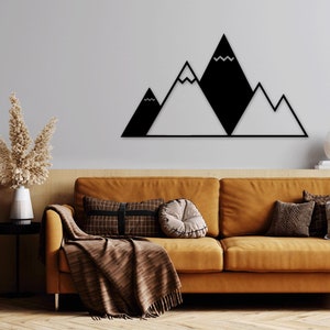 May include: Black metal wall art depicting a mountain range with three peaks. The mountains are stylized with a simple geometric design. The art is mounted on a white wall above a brown couch with pillows and a throw blanket.