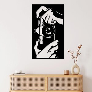 May include: Black and white silhouette wall art of hands holding a vintage camera. The camera has a large lens and a film rewind knob. The art is perfect for a home office or living room.