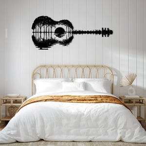 May include: Black metal wall art depicting a guitar silhouette with a sunset and trees reflected in water. The art is displayed above a white bed with a brown throw blanket and a wooden headboard.