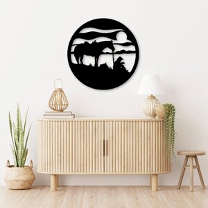 May include: Black metal wall art depicting a horse and rider silhouetted against a sunset with a full moon. The art is mounted on a white wall above a wooden cabinet and a small wooden stool.