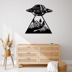 May include: Black metal wall art depicting a UFO hovering above a mountain range with trees. The artwork is mounted on a white wall above a wooden dresser with woven drawer fronts. The overall theme is extraterrestrial and nature.