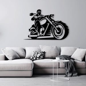 Op de afbeelding: Zwarte silhouet van een motorrijder op een motorfiets met een grote voorband. De afbeelding is op een witte muur boven een grijze bank met kussens en een bijzettafel.