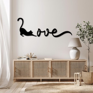 May include: Black metal wall decor featuring a cat silhouette and the word "love" in a cursive font. The decor is mounted on a white wall above a light brown wooden cabinet with woven rattan doors.